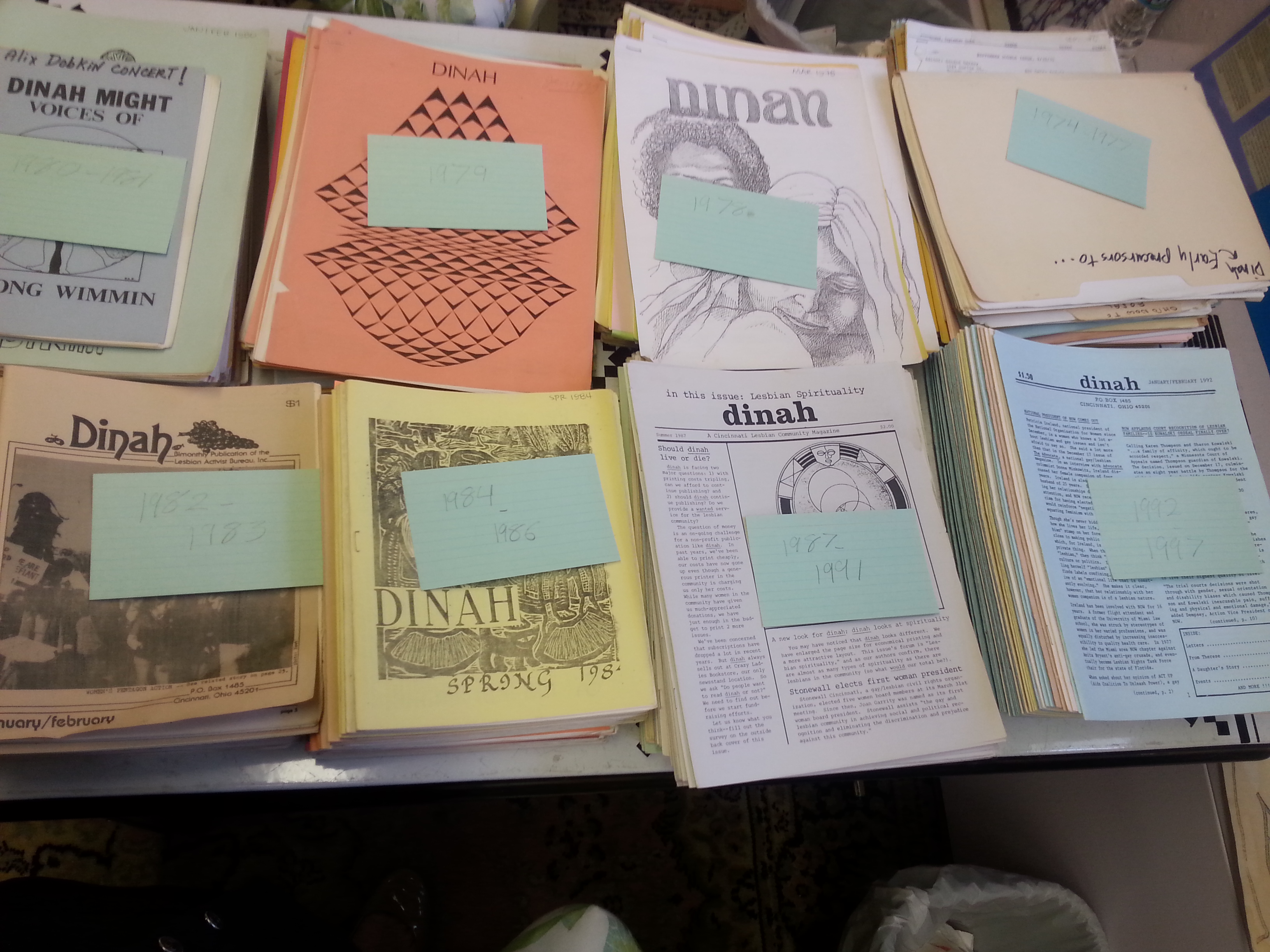 Photograph of eight piles of a multi-colored newsletter sitting on a table, each with an index card indicating a date range between 1975 and 1997.