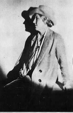 Black and white photograph of Mary MacMillan, facing left of frame, wearing hat, loose suit jacket, and a long necklave which she is holding in her right hand.